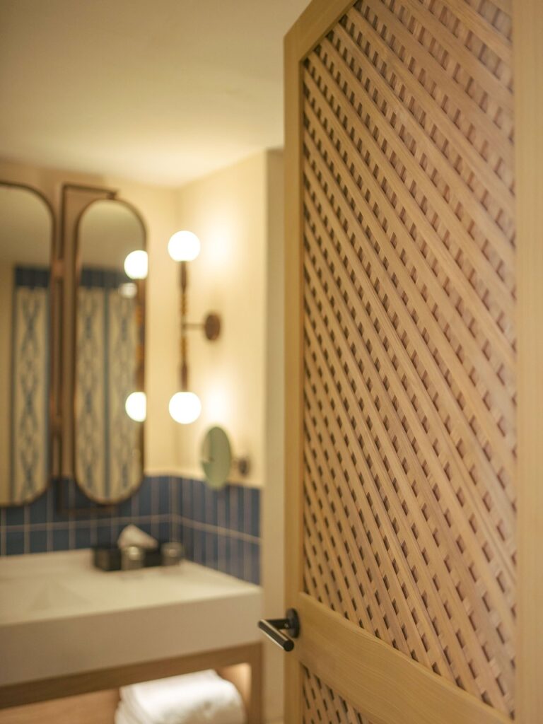 Puerta de celosía de madera abierta hacia el baño con mueble de lavabo e iluminación cálida