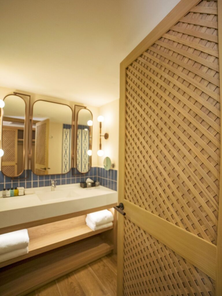 Baño de habitación en Ocean Coral & Turquesa con puerta de celosía de madera, mueble de lavabo y espejos de marco curvo